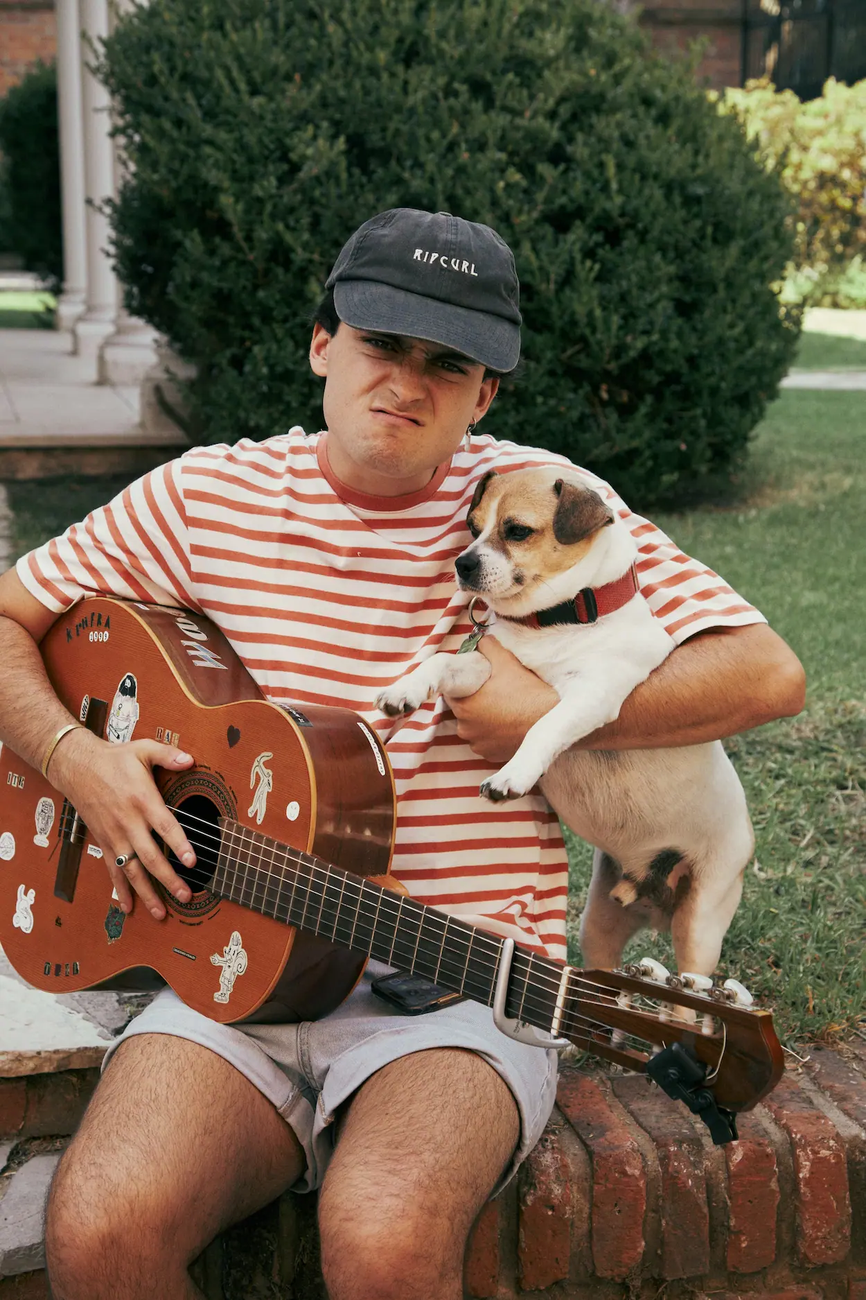 A man sitting on a brick wall holding a guitar and a dog