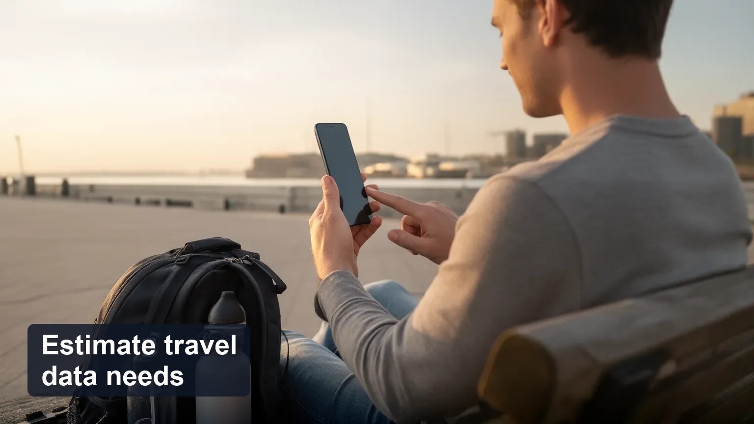 Traveler at a city viewpoint holding a phone with the screen turned away, with the city skyline in warm late-afternoon light.